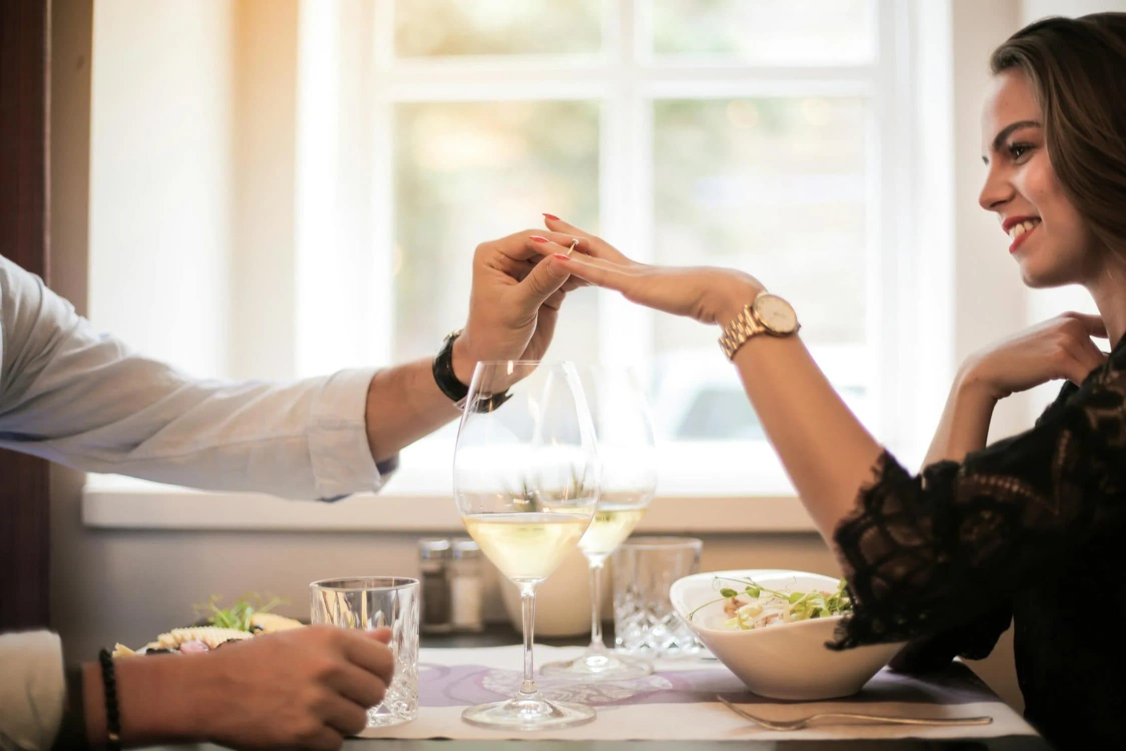 Man sliding an engagement ring onto a smiling woman's finger at a restaurant dinner