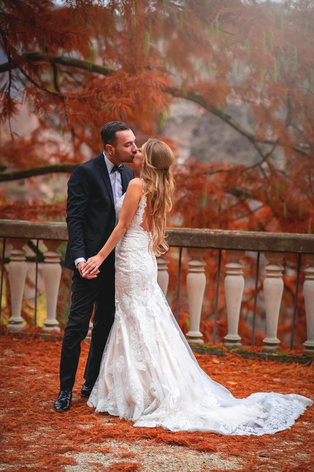 Bride and groom sharing a kiss beside a stone balustrade amid autumn foliage