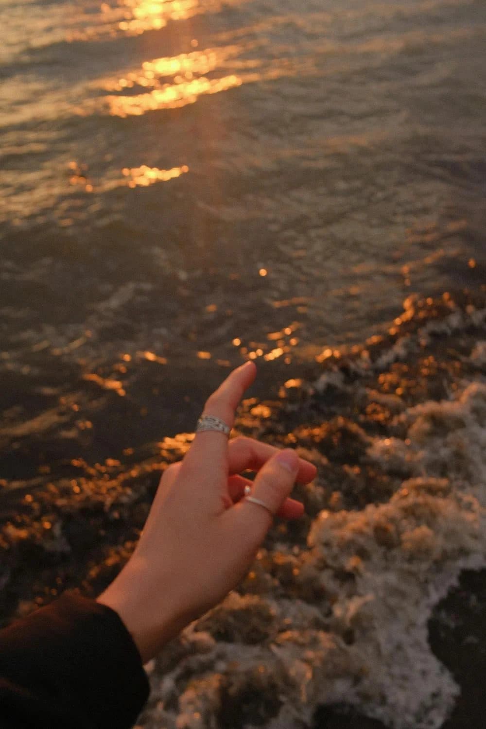 Hand wearing two silver rings raised above golden sunset ocean waves