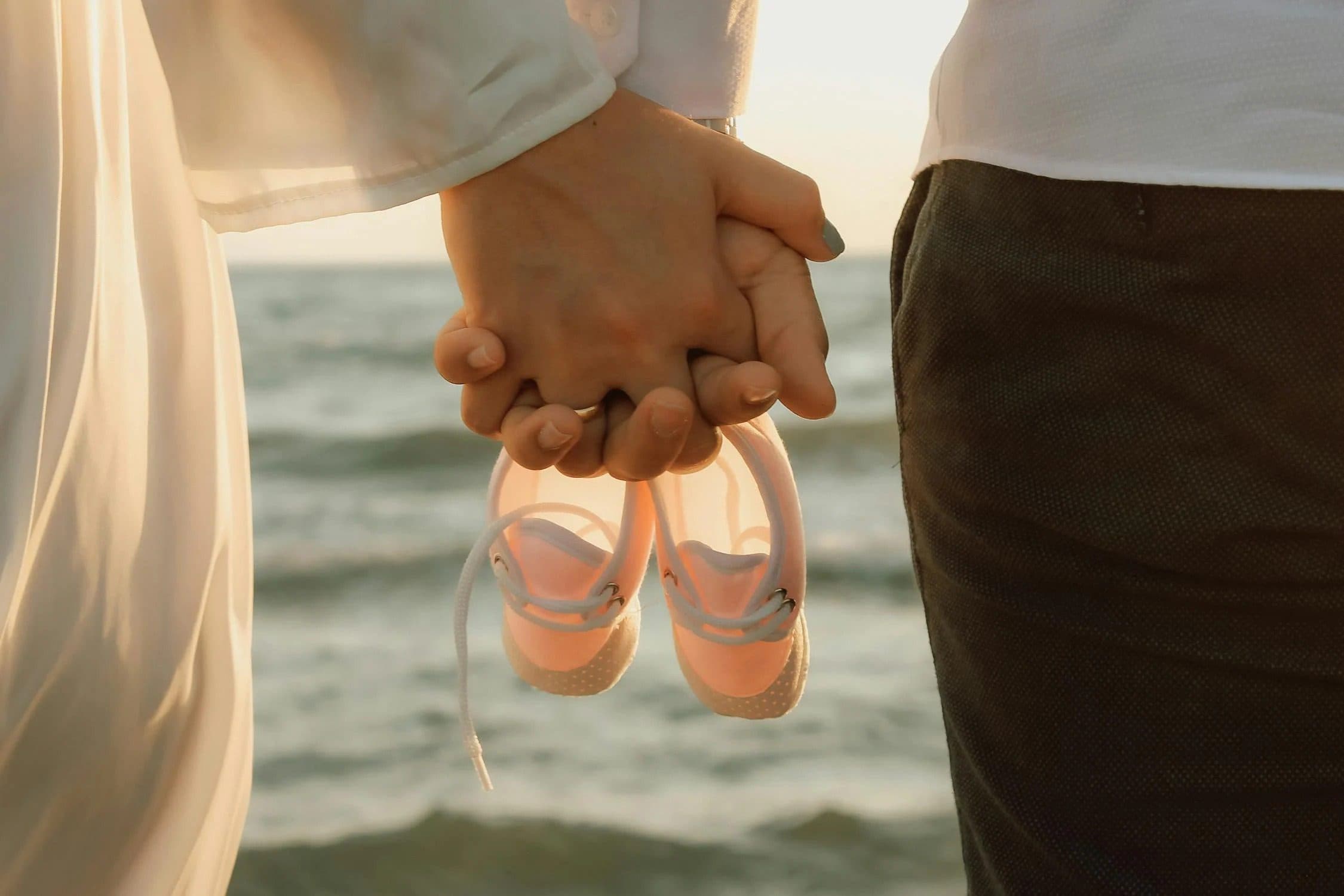 Couple holding hands and dangling tiny pink baby shoes at the beach