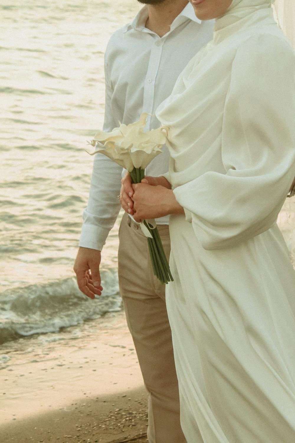 Bride in flowing white dress holding calla lily bouquet beside groom at shoreline