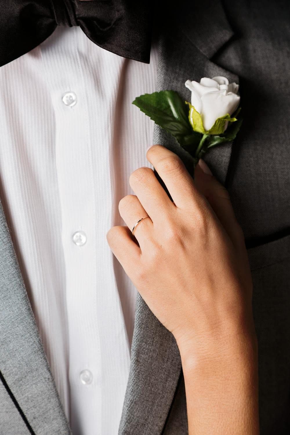 Hand wearing a slim silver wedding band pinning a white rose boutonniere on a tuxedo lapel