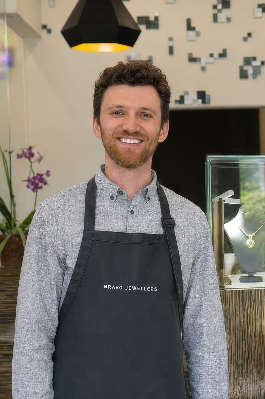 Smiling male jeweller in a Bravo Jewellers apron standing beside a display case