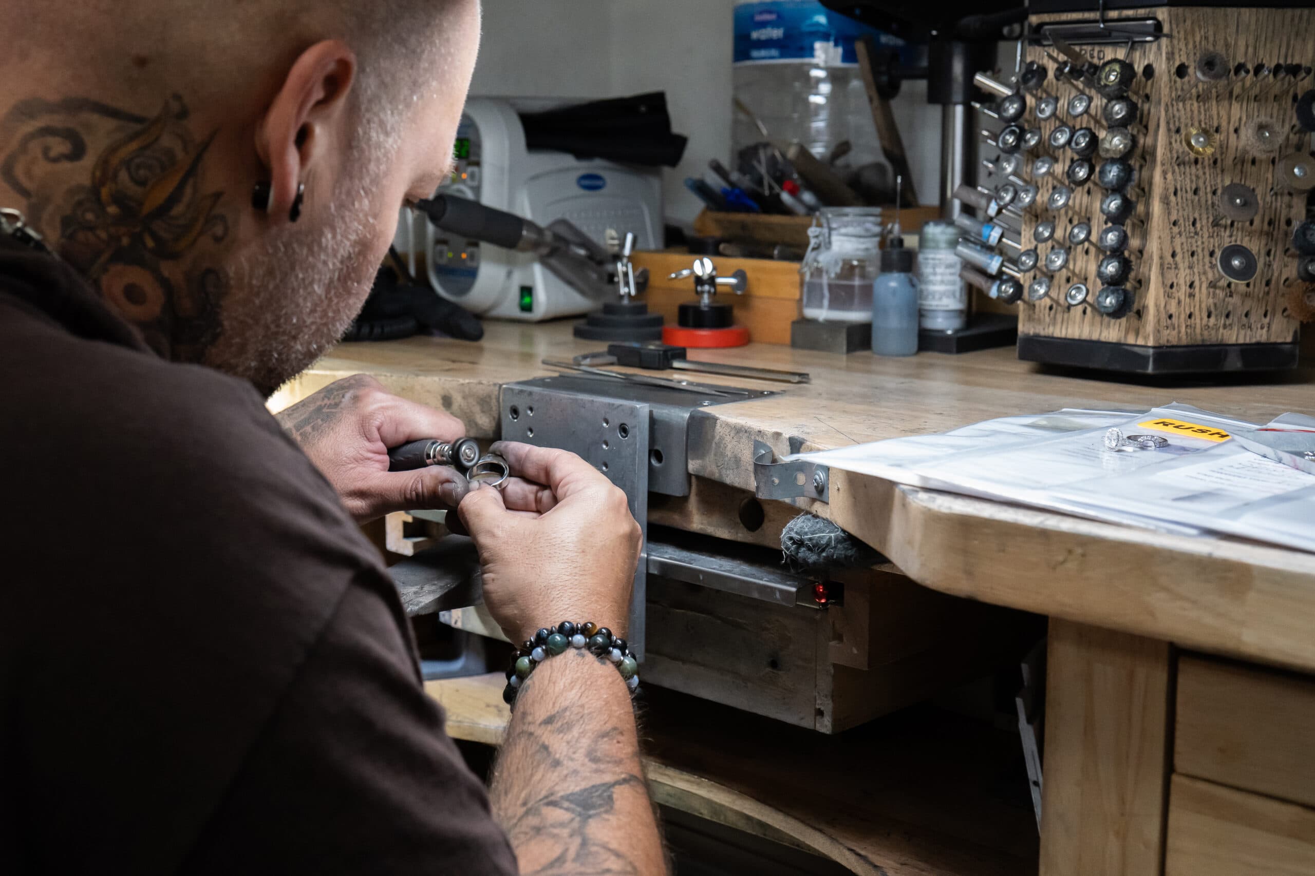 Tattooed jeweler using a hand tool to work on a small ring at a workshop bench
