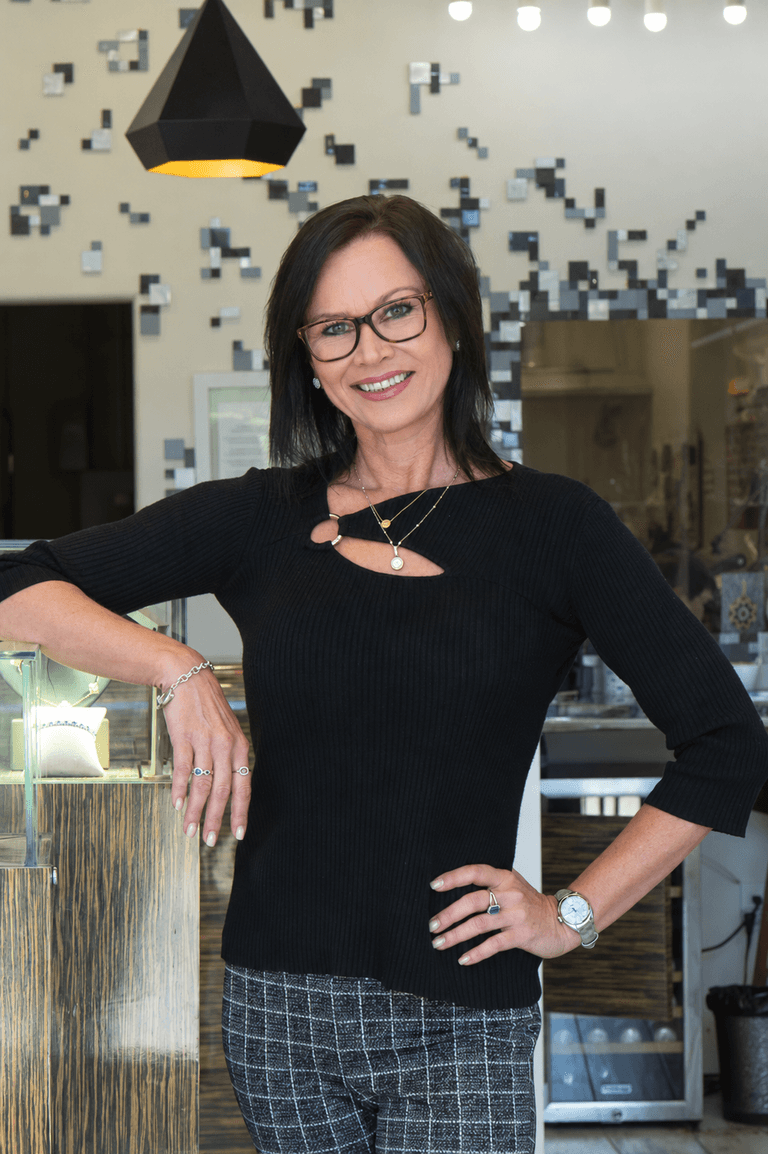 Smiling woman wearing layered gold necklaces, rings, and a silver watch beside a jewelry display case