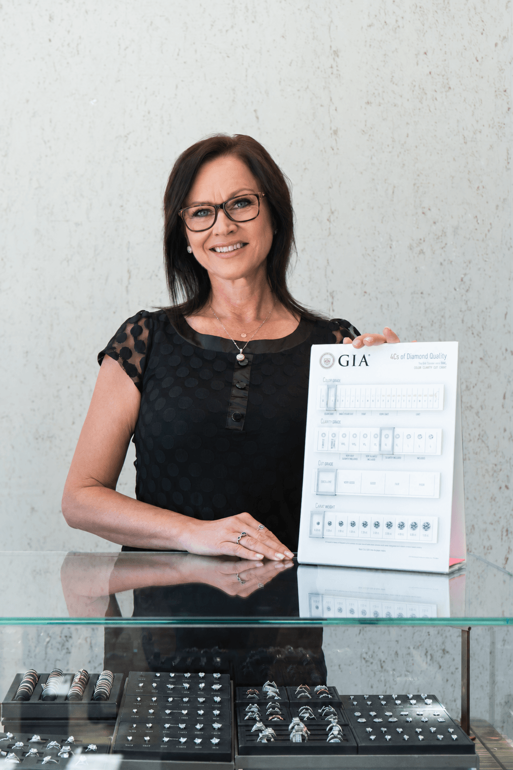 Smiling female jewelry consultant holding a GIA 4Cs of Diamond Quality display card at a glass showcase counter