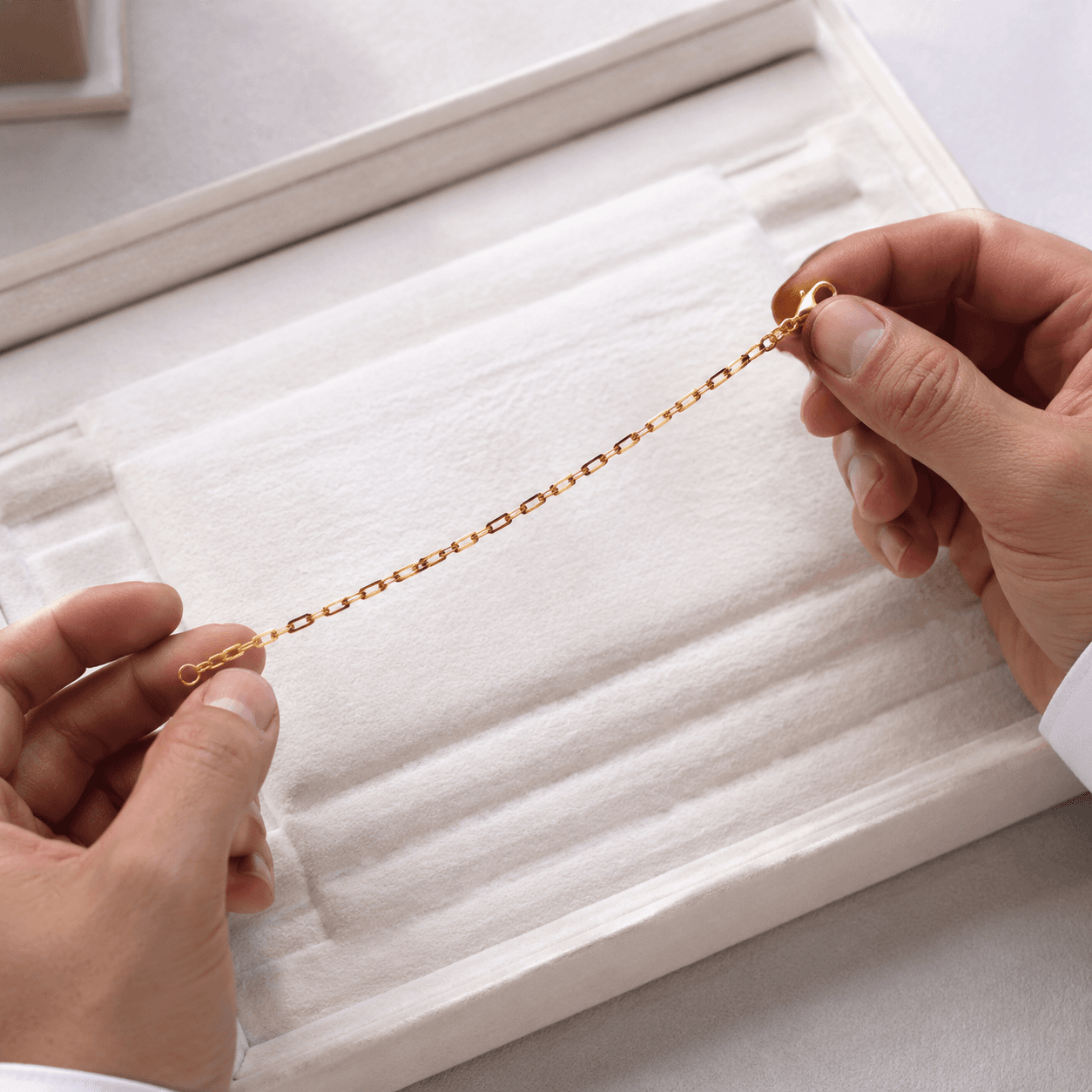 A jeweler's hands holding a delicate gold chain bracelet against a white velvet sizing board, overhead shot, soft studio