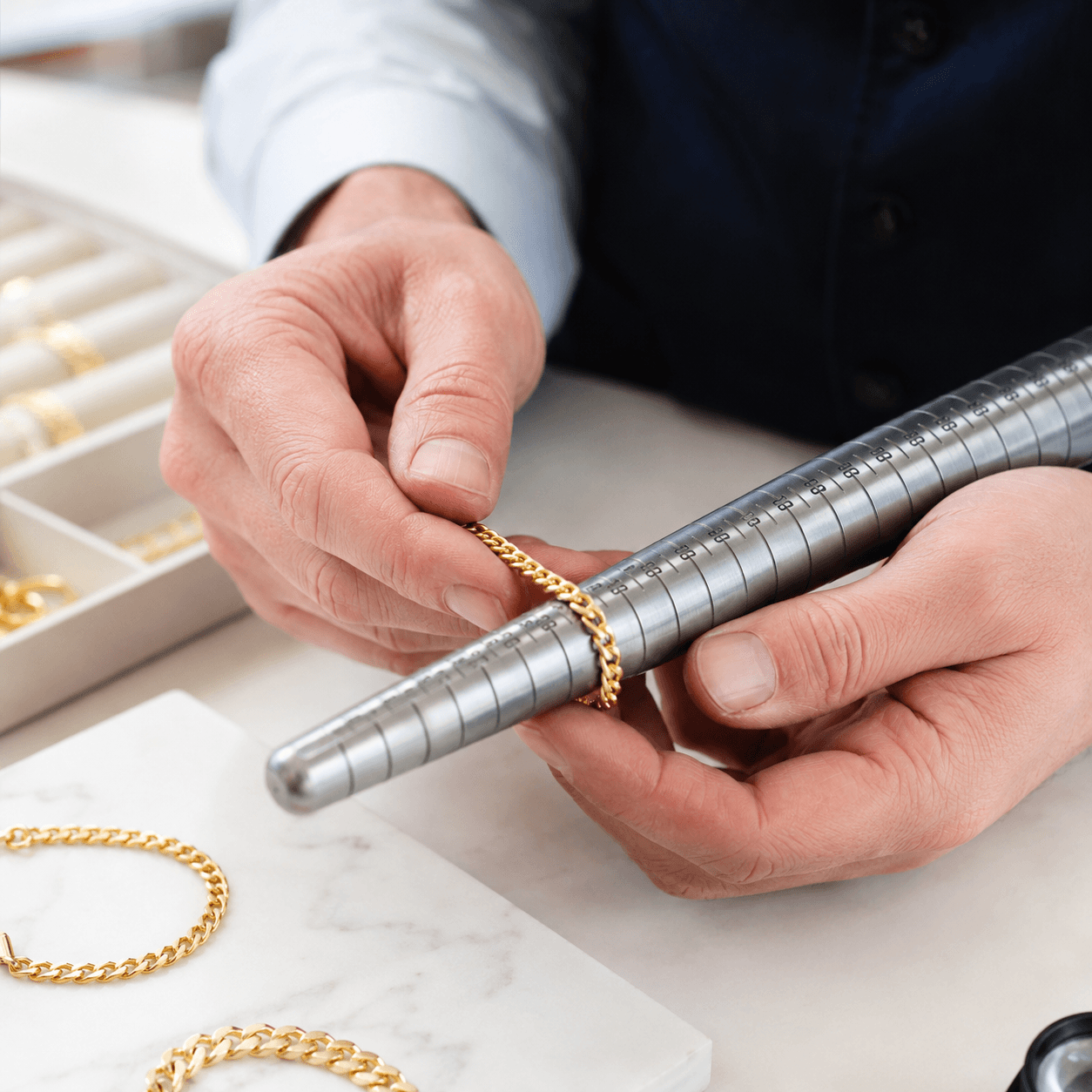 A jeweler's hands measuring a gold chain bracelet against a sizing gauge on a clean white workbench, close-up shot, bright