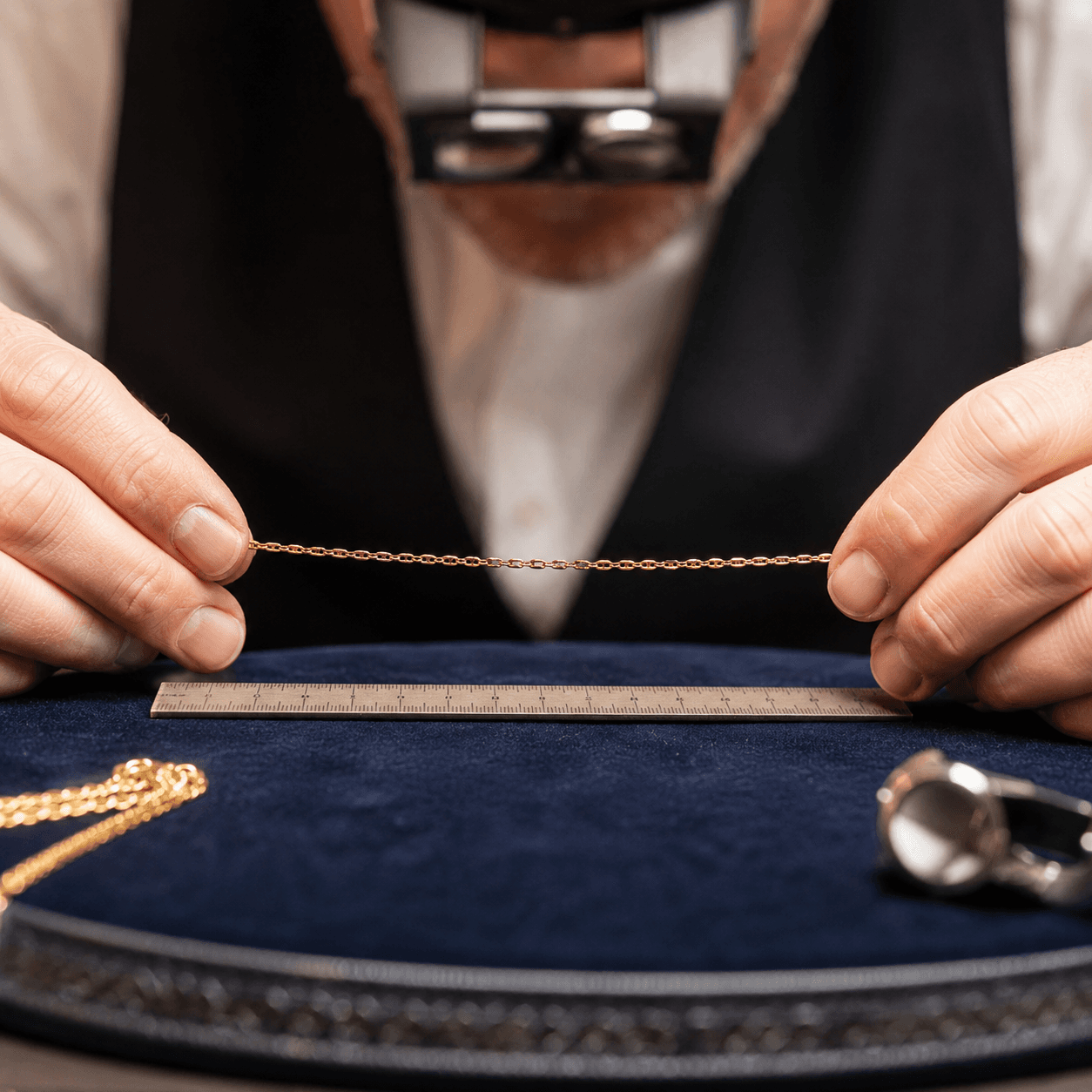 A jeweler holding a delicate gold chain necklace up to a measuring ruler on a velvet-lined workbench, close-up shot, warm