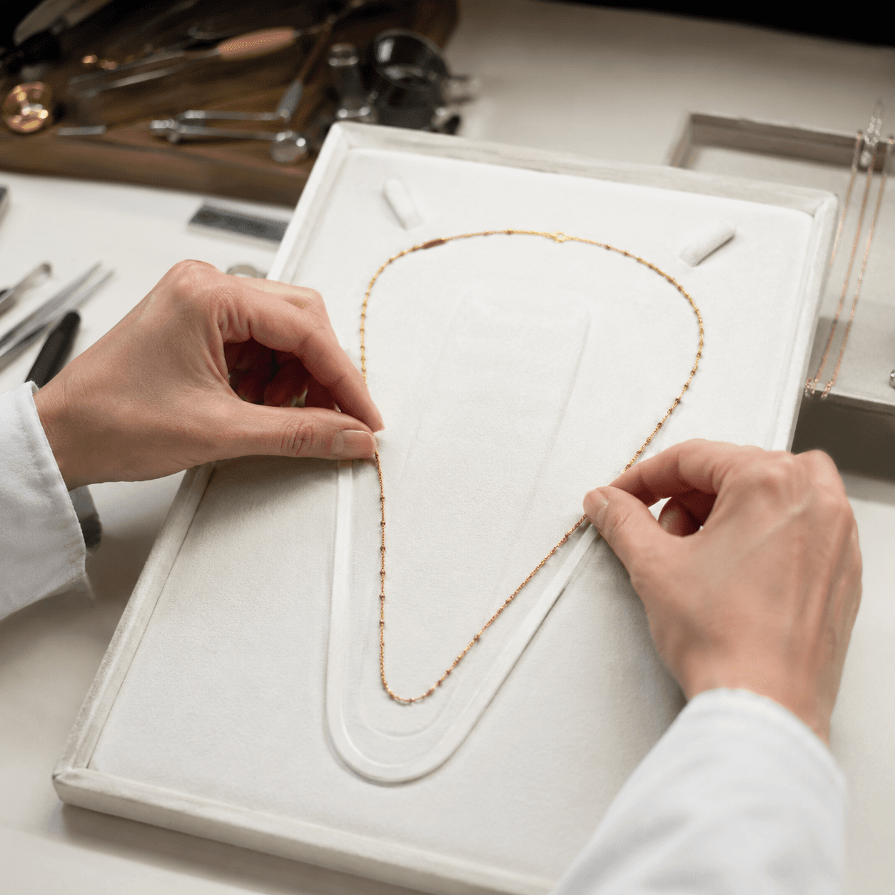 A jeweler's hands carefully measuring a delicate gold chain necklace against a white velvet sizing board on a clean workshop